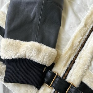 May include: Close-up of a black leather jacket with cream-colored faux fur lining and trim. The jacket features a black ribbed knit cuff, a black leather strap with a gold-colored buckle, and a gold-colored zipper.