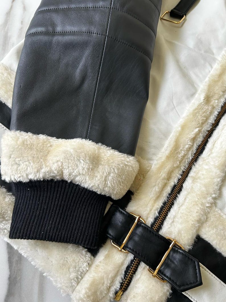 May include: Close-up of a black leather jacket with cream-colored faux fur lining and trim. The jacket features a black ribbed knit cuff, a black leather strap with a gold-colored buckle, and a gold-colored zipper.