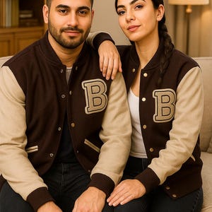 May include: A couple wearing matching brown and beige letterman jackets, each with a large "B" on the chest. The jackets have a button-up front and ribbed cuffs. They are sitting on a beige sofa.