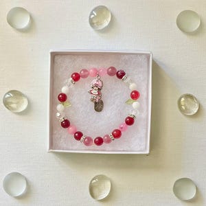 Peut inclure: Un bracelet de perles roses, rouges et blanches, agrémenté d'entretoises argentées et d'un charm. Le charm représente un personnage de dessin animé et l'inscription "MADE WITH LOVE". Le bracelet est présenté dans une boîte blanche.