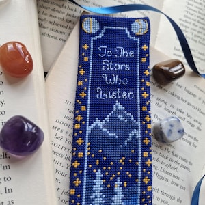 May include: A rectangular blue cross-stitch bookmark featuring the white text "To The Stars Who Listen" above a mountain landscape with yellow stars. The bookmark is accented with a blue ribbon and surrounded by polished orange, brown, and blue stones.
