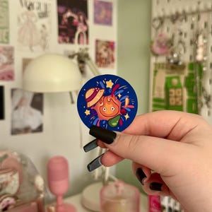 May include: A circular navy blue sticker with a cartoon character design. The character has orange flames, a straw hat, and stars. The sticker is held in a hand with black nail polish, with a blurred background of wall art and a pink microphone.