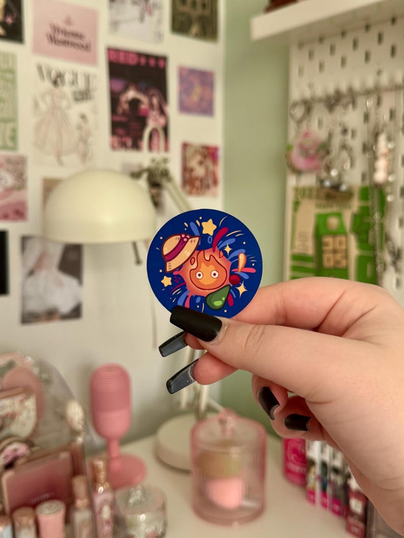 May include: A circular navy blue sticker with a cartoon character design. The character has orange flames, a straw hat, and stars. The sticker is held in a hand with black nail polish, with a blurred background of wall art and a pink microphone.