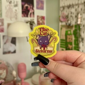May include: A yellow sticker featuring the text "The Cat Returns" and a purple cat illustration. The sticker has a decorative border. The sticker is held up by a person.
