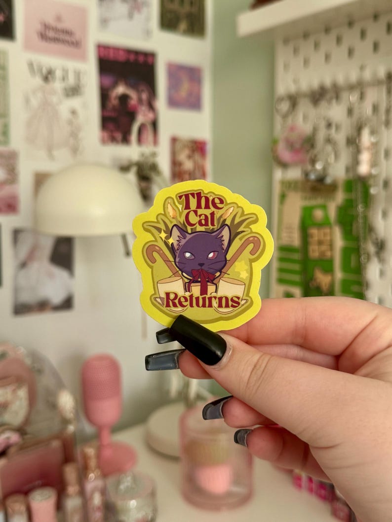 May include: A yellow sticker featuring the text "The Cat Returns" and a purple cat illustration. The sticker has a decorative border. The sticker is held up by a person.