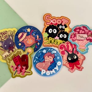 May include: A selection of vibrant stickers showcasing characters and scenes from animated films. Designs include a black cat, a fiery character, soot sprites, a bouquet of flowers, and a character with fish. The stickers are on a white and green background.