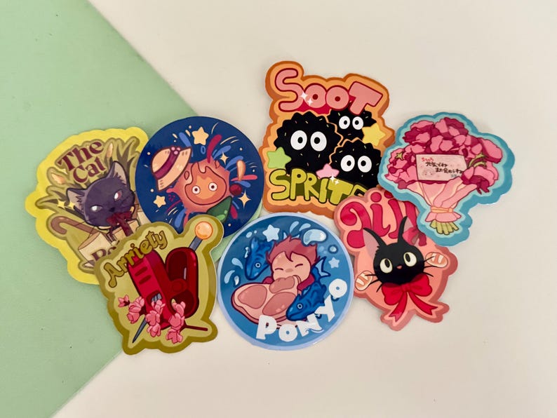 May include: A selection of vibrant stickers showcasing characters and scenes from animated films. Designs include a black cat, a fiery character, soot sprites, a bouquet of flowers, and a character with fish. The stickers are on a white and green background.