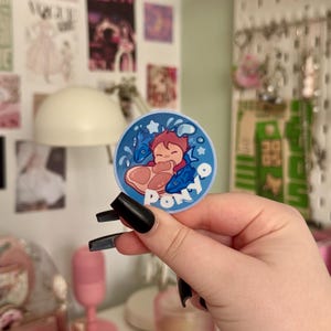 May include: A circular sticker with a cartoon illustration of a character with red hair and two blue fish. The sticker has a blue background with white stars and the word "Ponyo" in white. The sticker is held by a person with black nail polish.