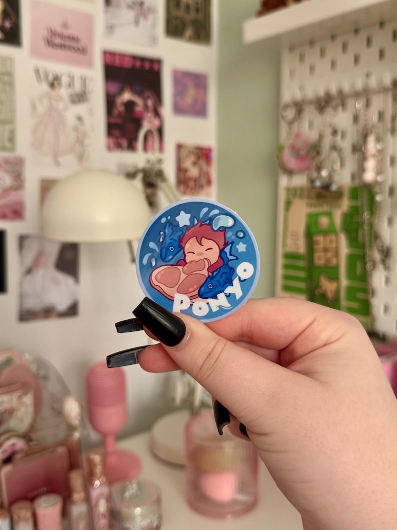 May include: A circular sticker with a cartoon illustration of a character with red hair and two blue fish. The sticker has a blue background with white stars and the word "Ponyo" in white. The sticker is held by a person with black nail polish.