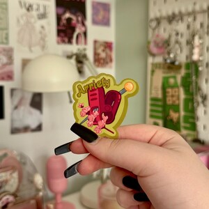 May include: A yellow sticker with the word "Arriety" in a stylized font. The sticker features a red heart, a sewing machine, and pink flowers. A needle with a yellow head is also present. The sticker is held up by a person.
