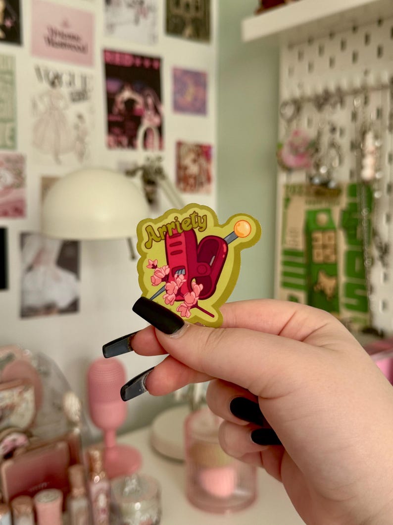 May include: A yellow sticker with the word "Arriety" in a stylized font. The sticker features a red heart, a sewing machine, and pink flowers. A needle with a yellow head is also present. The sticker is held up by a person.