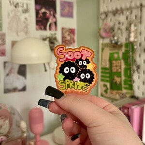 May include: A colourful sticker with the text "Soot Sprite" and a cartoon design of three black, round characters with white eyes. The sticker has an orange, black, and green colour scheme and is held by a hand with black nail polish.
