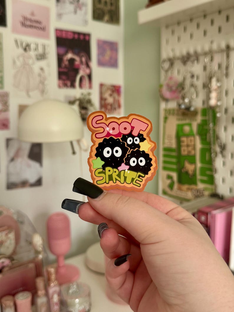 May include: A colourful sticker with the text "Soot Sprite" and a cartoon design of three black, round characters with white eyes. The sticker has an orange, black, and green colour scheme and is held by a hand with black nail polish.