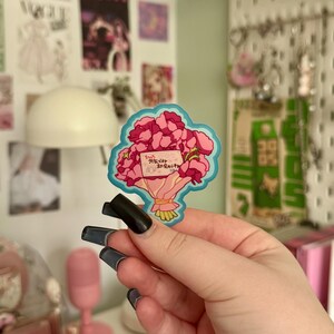 May include: A sticker depicting a bouquet of pink flowers with a blue outline. The bouquet is tied with a yellow ribbon and includes a small card with Japanese text. The sticker is held in a hand with long, dark nails.