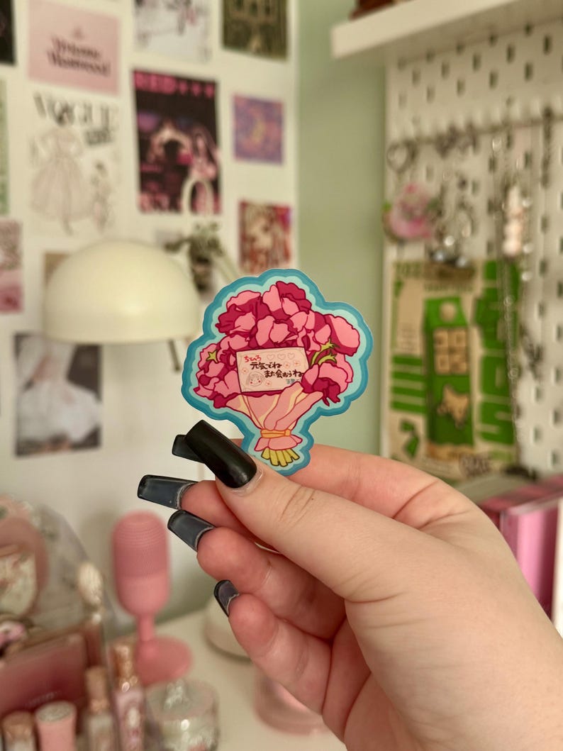 May include: A sticker depicting a bouquet of pink flowers with a blue outline. The bouquet is tied with a yellow ribbon and includes a small card with Japanese text. The sticker is held in a hand with long, dark nails.