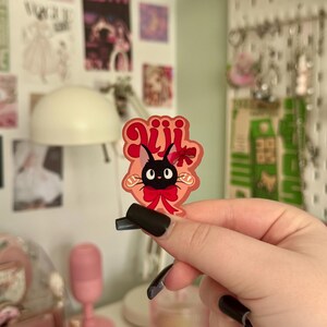 May include: A sticker featuring a black cat with large eyes, a red bow, and the Japanese text "Jiji". The sticker has a pink and red border. The cat is a character from the animated film Kiki's Delivery Service.