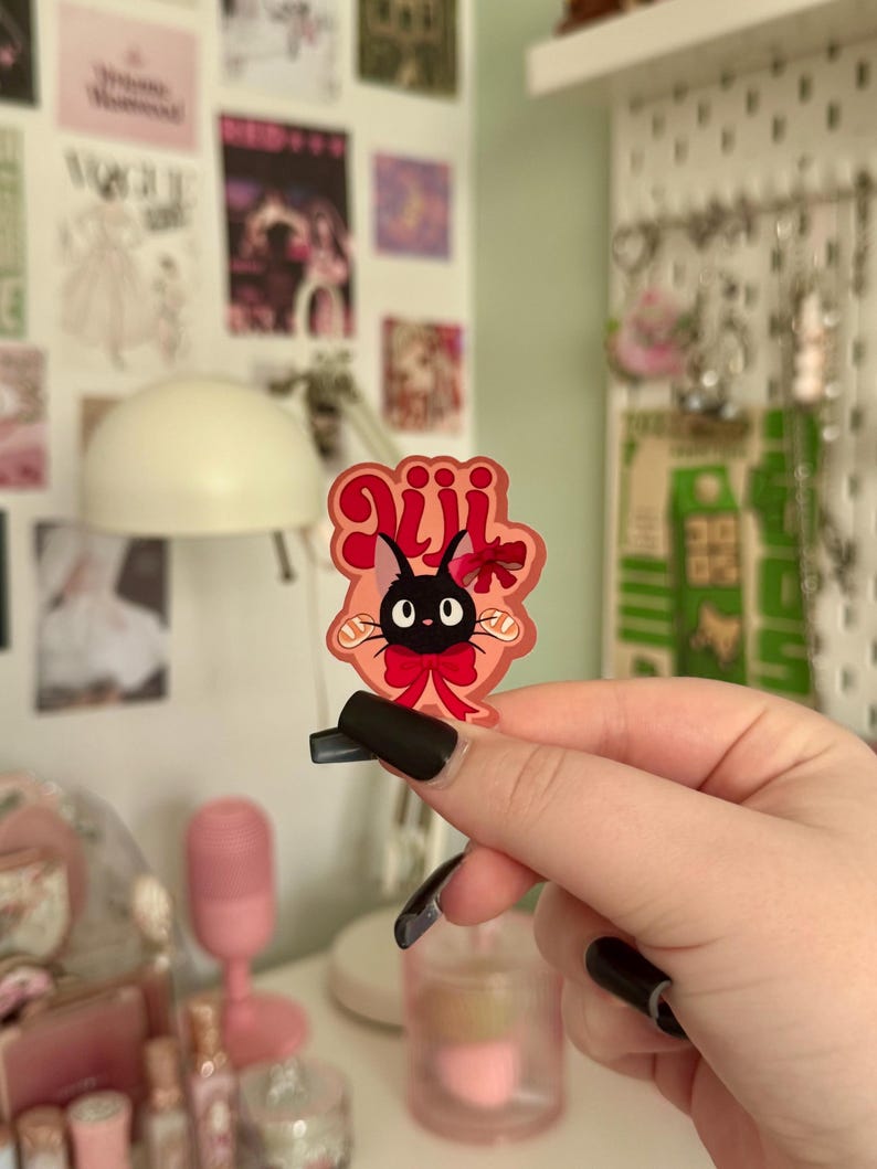 May include: A sticker featuring a black cat with large eyes, a red bow, and the Japanese text "Jiji". The sticker has a pink and red border. The cat is a character from the animated film Kiki's Delivery Service.