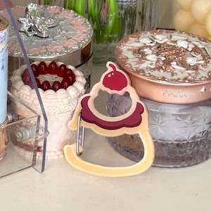 May include: A decorative mirror in the shape of a cupcake with a cherry on top. The mirror has a yellow base and a burgundy and white trim. Other decorative items are in the background.