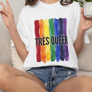 May include: White t-shirt with a rainbow watercolor design and the text "TRÈS QUEER" in black. The design features vertical brushstrokes in red, orange, yellow, green, blue, and purple. The shirt is a casual style.