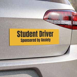 Peut inclure: Un autocollant de pare-chocs rectangulaire jaune avec le texte "Student Driver Sponsored by Anxiety" en lettres noires est apposé à l'arrière d'une voiture argentée. Le feu arrière de la voiture est visible.