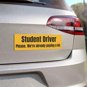 Op de afbeelding: Een gele rechthoekige bumpersticker met zwarte tekst: "Student Driver Please. We're already paying a lot." De sticker is bevestigd aan de achterkant van een zilveren auto.