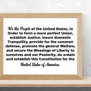 May include: A framed print of the Preamble to the United States Constitution. The text is in black font on a white background. The frame is a light wood colour.