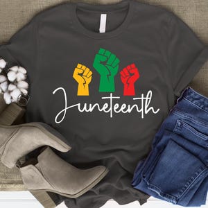 May include: Dark gray t-shirt with the word "Juneteenth" in white script. The shirt features three raised fists in yellow, green, and red. A pair of gray ankle boots and blue jeans are also in the image.