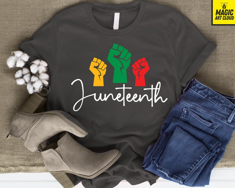 May include: Dark gray t-shirt with the word "Juneteenth" in white script. The shirt features three raised fists in yellow, green, and red. A pair of gray ankle boots and blue jeans are also in the image.