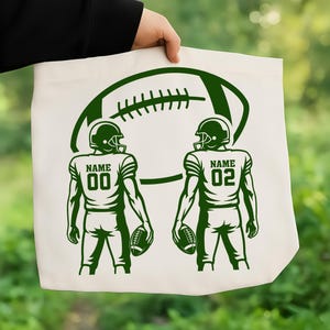 Puede incluir: Un bolso tote de color crema con un gr&aacute;fico verde de un bal&oacute;n de f&uacute;tbol americano y dos jugadores. Cada jugador lleva un casco y sostiene un bal&oacute;n. El texto "NAME" y un n&uacute;mero est&aacute;n impresos en las camisetas.