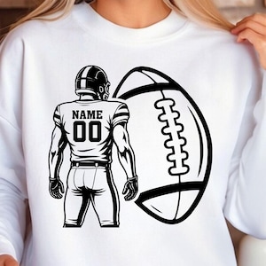Puede incluir: Sudadera blanca con un gr&aacute;fico en blanco y negro de un jugador de f&uacute;tbol americano y un bal&oacute;n. La camiseta del jugador muestra "NAME" sobre el n&uacute;mero "00". El bal&oacute;n tambi&eacute;n es blanco y negro. Dise&ntilde;o gr&aacute;fico audaz.