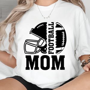 Puede incluir: Camiseta blanca con un dise&ntilde;o de casco y bal&oacute;n de f&uacute;tbol en blanco y negro. Las palabras "FOOTBALL MOM" est&aacute;n impresas en letras negras en negrita. Un gran regalo para las madres futbolistas.
