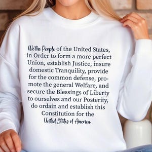 May include: White sweatshirt with the text "We the People of the United States, in Order to form a more perfect Union, establish Justice, insure domestic Tranquility, provide for the common defence, promote the general Welfare, and secure the Blessings of Liberty to ourselves and our Posterity, do ordain and establish this Constitution for the United States of America."
