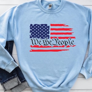 May include: Light blue sweatshirt featuring a distressed American flag design and the text "We the People" in black.