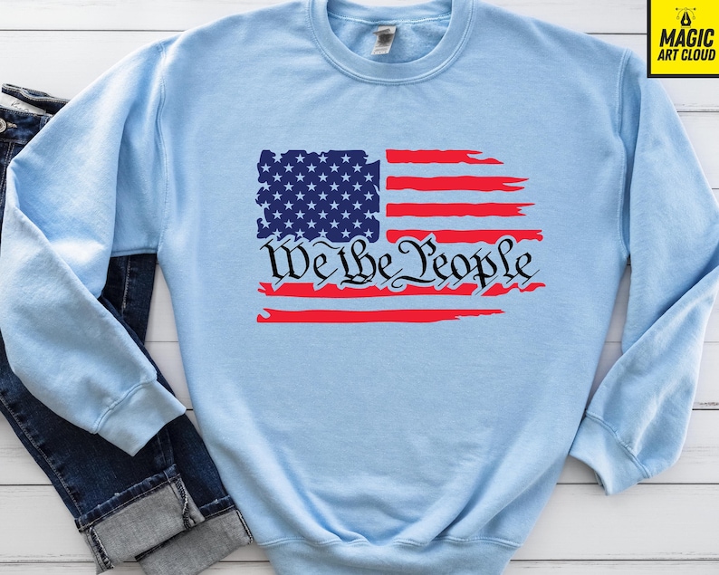 May include: Light blue sweatshirt featuring a distressed American flag design and the text "We the People" in black.