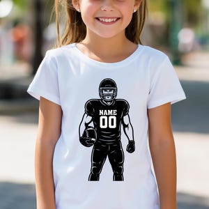 Puede incluir: Camiseta blanca con una ilustraci&oacute;n en blanco y negro de un jugador de f&uacute;tbol americano sosteniendo un bal&oacute;n. La camiseta del jugador muestra el nombre y el n&uacute;mero "00". La camiseta la lleva una persona.