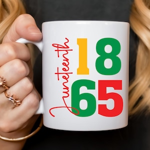 May include: White ceramic mug featuring the word "Juneteenth" in red script, and the numbers "18" in yellow and green, and "65" in red. The mug has a white handle and is held by a person.