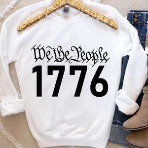 May include: White sweatshirt with black text that reads "We the People 1776".