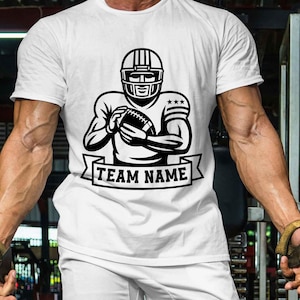 Puede incluir: Camiseta blanca con una ilustraci&oacute;n en blanco y negro de un jugador de f&uacute;tbol americano con casco, sosteniendo un bal&oacute;n. El dise&ntilde;o incluye una pancarta con el texto "TEAM NAME".
