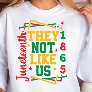 May include: White sweatshirt with a colorful graphic design. The design features the text "THEY NOT LIKE US" in yellow and green, with the word "Juneteenth" in red. The design also includes stars and the numbers 1, 8, 6, and 5.
