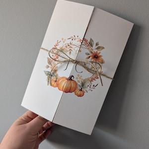 May include: A folded white card with a watercolour design of a wreath with pumpkins, leaves, and sunflowers. The wreath is tied with twine and the initials "J C" are in the centre.