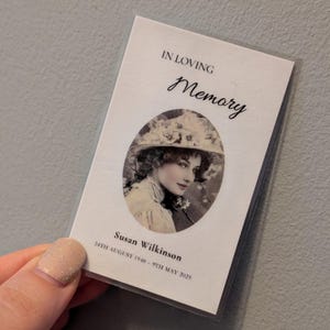 May include: A white memorial card with the words "IN LOVING Memory" in script. It features a black and white portrait of a woman in a hat. The name "Susan Wilkinson" and dates are printed below the portrait.