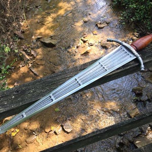 May include: A medieval-style sword with a silver blade and a brown wooden handle. The sword is resting on a wooden surface, with a stream of water and rocks in the background. The sword has a decorative hilt and a pointed tip.