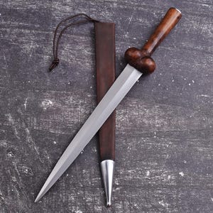 May include: A dagger with a polished steel blade and a dark wooden handle. The handle has a bulbous design. The dagger rests in a brown leather sheath with a silver tip. A leather cord is attached to the sheath.