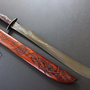 May include: A detailed image of a sword and its sheath. The sword has a silver blade with etched designs and a wooden handle. The sheath is brown with carved patterns and text. A small feather and bead dangle from the sheath.