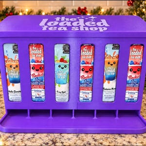 Puede incluir: Un estante dispensador de plástico morado con la etiqueta "the loaded tea shop" en la parte superior, que cuenta con seis ranuras verticales que contienen paquetes de mezcla para bebidas. Cada paquete muestra un personaje y texto.