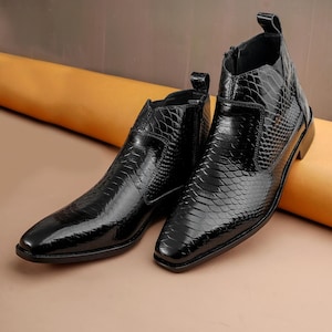 May include: A pair of black leather ankle boots with a textured, reptile-like pattern. The boots have a pointed toe, a side zipper, and a pull tab at the back. The boots are glossy and reflect light.
