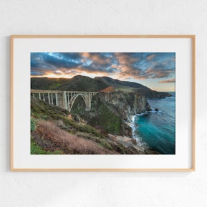 May include: A framed print of Bixby Bridge on the California coast. The concrete arch bridge spans a deep canyon, with the Pacific Ocean visible below. The sky is filled with colourful clouds at sunset, and the surrounding cliffs are covered in green vegetation.