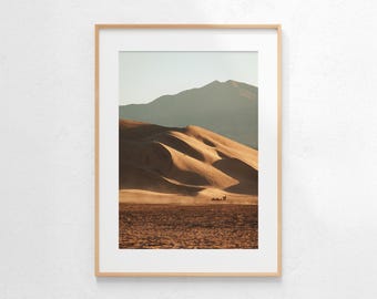 May include: Framed landscape photograph featuring a desert scene with sand dunes and a mountain in the background. The dunes are a warm brown color, and the sky is a pale blue. The photograph is in a light wood frame.