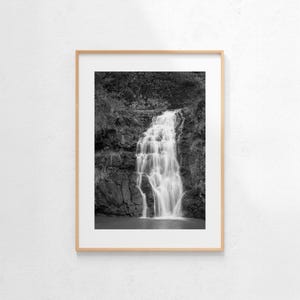 May include: Black and white framed photograph of a waterfall cascading over layered rocks. The water flows down in multiple tiers, surrounded by dark foliage. The photograph is in a wooden frame.