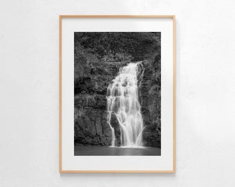 May include: Black and white framed photograph of a waterfall cascading over layered rocks. The water flows down in multiple tiers, surrounded by dark foliage. The photograph is in a wooden frame.